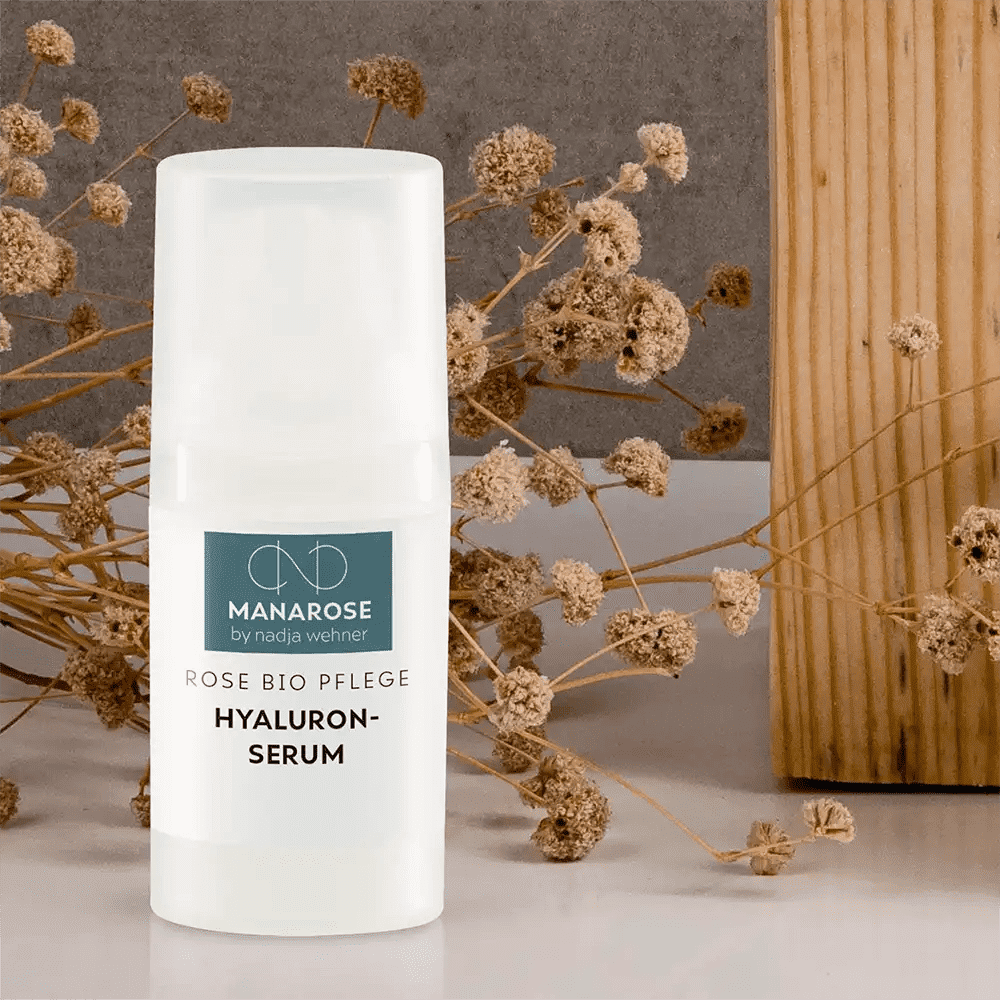 Eine Flasche Manarose Biokosmetik Hyaluron Serum Rose versorgt die Haut mit Feuchtigkeit, neben einer Schachtel Trockenblumen.