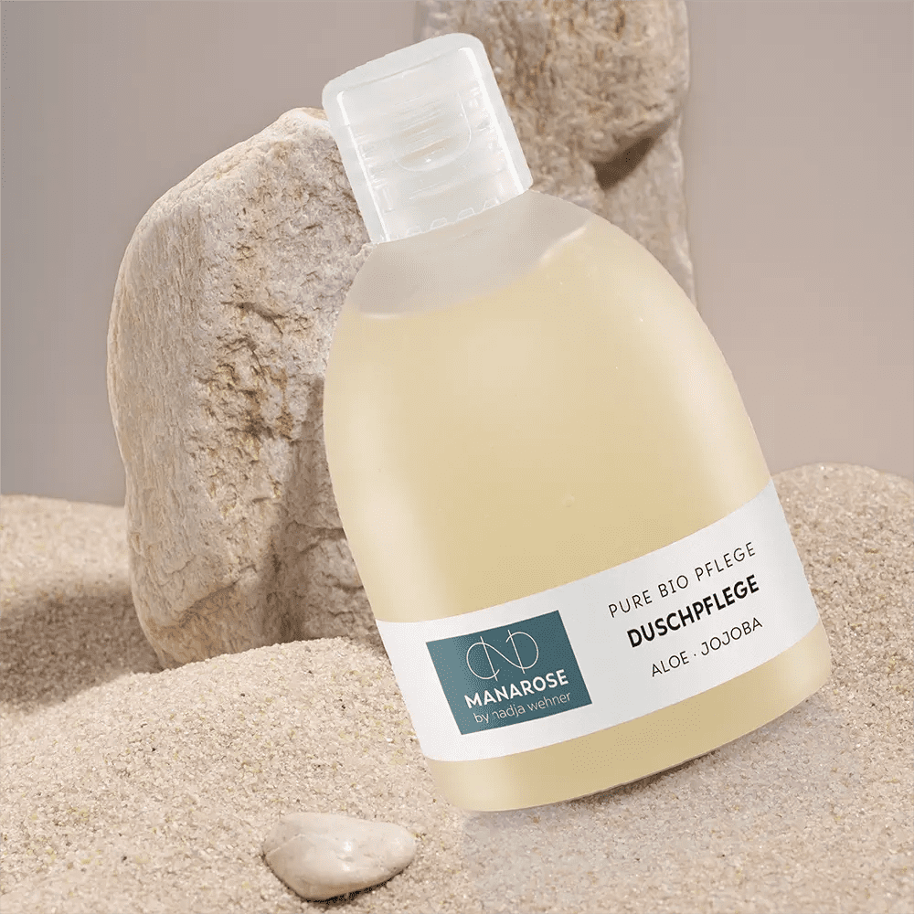 Eine Flasche Duschpflege Aloe Jojoba von Manarose Biokosmetik liegt neben Felsen im Sand.