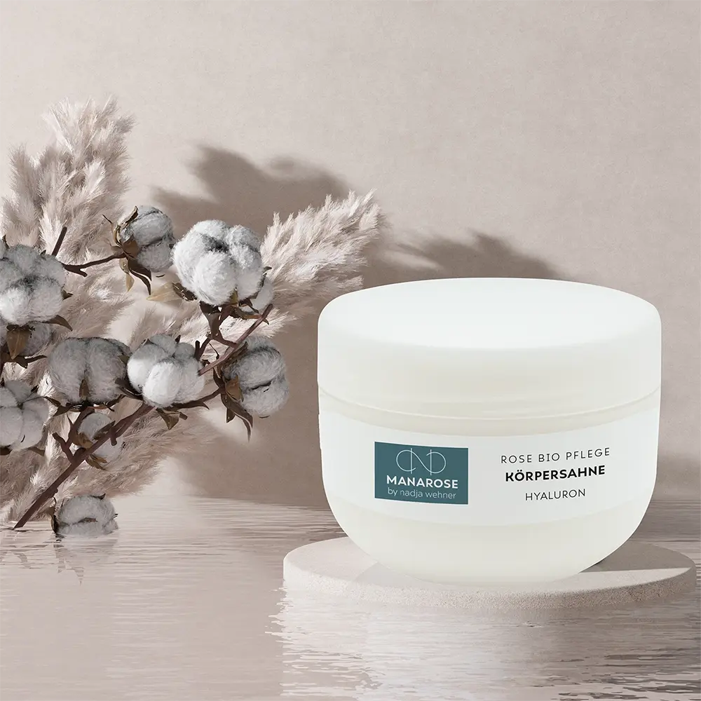 Ein weißes Glas mit einer Baumwollpflanze und darauf Körperahne Rose Hyaluron von Manarose Biokosmetik.