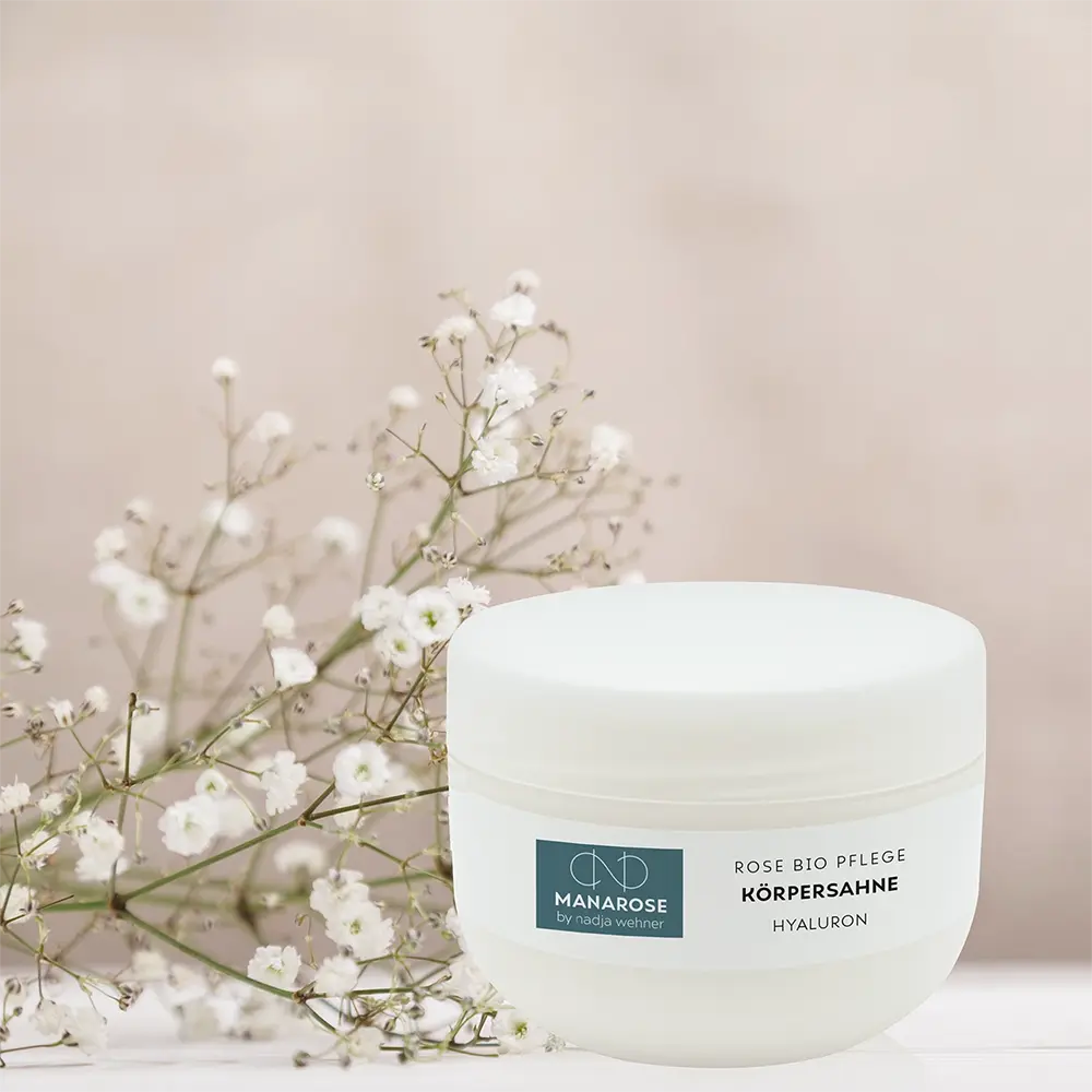 Ein Glas feuchtigkeitsspendendes körperähnliches Rosen-Hyaluron von Manarose Biokosmetik auf einem weißen Tisch mit Blumen, der für ein pflegendes Hautgefühl und einen dezenten Rosenduft sorgt.