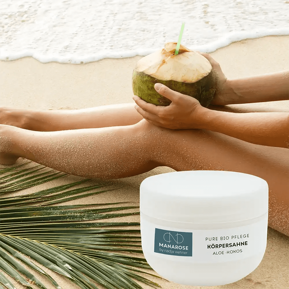 Eine Frau sitzt mit einer Kokosnuss in der Hand am Strand und trägt Körperahne Aloe Kokos von Manarose Biokosmetik auf ihre Haut auf, die sie pflegt und nährt.