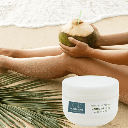 Eine Frau sitzt mit einer Kokosnuss in der Hand am Strand und trägt Körperahne Aloe Kokos von Manarose Biokosmetik auf ihre Haut auf, die sie pflegt und nährt.