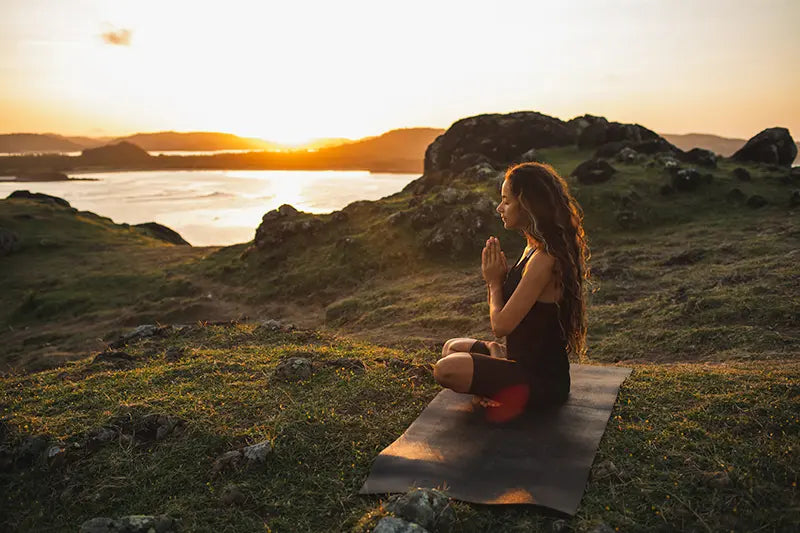 Eine Frau meditiert in der Natur hinter ihr sieht man den Sonnenuntergang